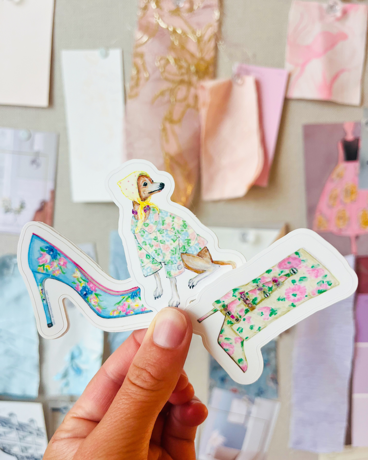 A hand holds two die-cut stickers featuring illustrated high-heeled shoes and a dog in a floral dress, with a soft-focus fashion board in the background.