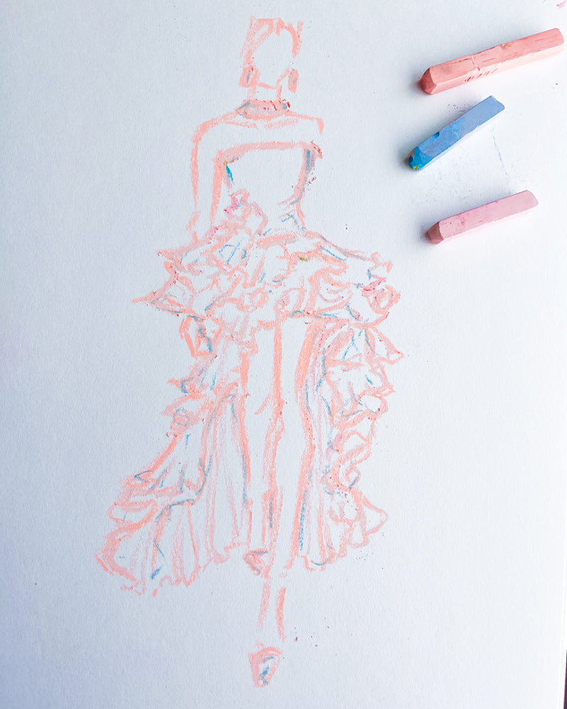 A fashion illustration sketch of a woman in a voluminous ruffled gown, drawn in peach-colored chalk on white paper, with three colored chalk sticks placed in the upper right corner.